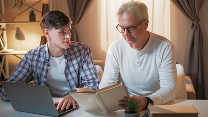 Homework help. Fatherhood leisure. Happy family. Cheerful man with book supporting son studying lesson together at laptop home interior.