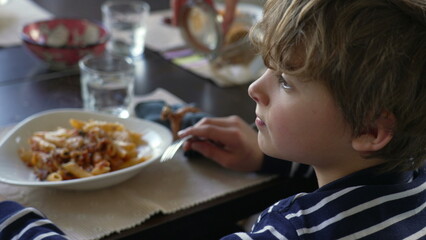 Pensive profile face of a small boy seated at lunch table with contemplative gaze. Child holding...