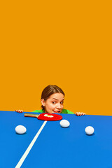 Woman peeks over table tennis table with paddle and balls, playful expression against orange...