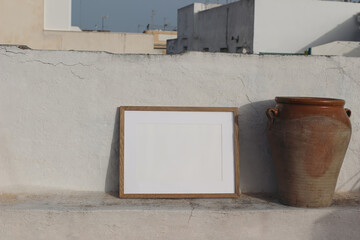 Blank horizontal wooden frame picture mock up. Vintage olive clay storage pot, vase. White old textured white wall in sunlight. Front view, no people. Summer display background for art, posters.