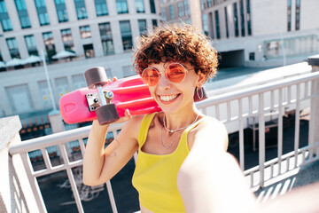 Young beautiful smiling hipster woman in trendy summer clothes. Carefree woman with curls hairstyle, posing in the street. Positive model outdoors. Cheerful and happy. Holds penny skateboard, selfie