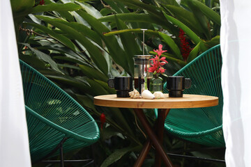 breakfast table with cups and French press, two acapulco chairs and plants in the background