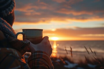 A person holding a coffee cup in front of a stunning sunset - obrazy, fototapety, plakaty