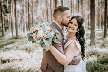 Valmiera, Latvia- July 28, 2024 - Groom embraces bride from behind in a forest, kissing her cheek;...