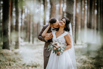 Valmiera, Latvia- July 28, 2024 - Groom kissing bride's cheek in the forest, bride holding a...