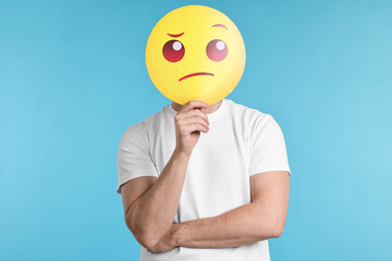 Man covering face with thinking emoticon on light blue background - obrazy, fototapety, plakaty