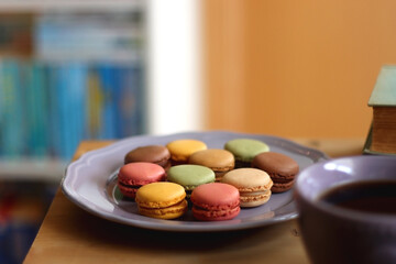 Purple plate filled with pastel macarons, cup of tea or coffee, vintage books and reading glasses on the table. Colorful bookcase in the background. Selective focus.