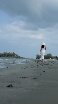 White dress Walking barefoot on the beach. European girl wearing Vietnamese hat. Beach Travel Woman walking on sandy beach, leaving footprints in sand. Close up detail of female legs and golden sand