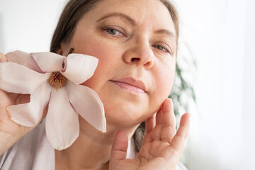 aging woman with magnolia flower looks herself in mirror, noticing wrinkles, changes in facial...
