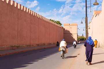 people walk along high wall Red City Marrakech hurry about business, authentic urban African...