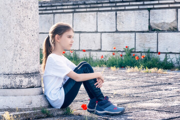 Portrait of a beautiful girl with a thoughtful look, seated at the base of a stone column and her hands resting on her knees.