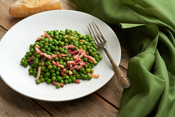 A white plate is filled with vibrant green peas and crispy pieces of bacon, accompanied by a silver fork on the table next to it.