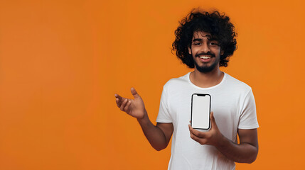 Positive curlyhaired happy Indian guy in white tshirt holds smartphone with mockup blank white screen stands on isolated orange background looks at camera and smiles friendly : Generative AI