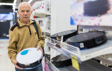 Elderly man choosing robot vacuum cleaner in showroom of electrical appliance store