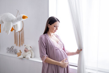 A pregnant woman strokes her big belly looking out the window in a bright children's bedroom by the...
