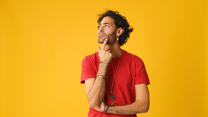 Pensive attractive man with curly hair, wearing red T-shirt,  man rubbing his chin and looking side...