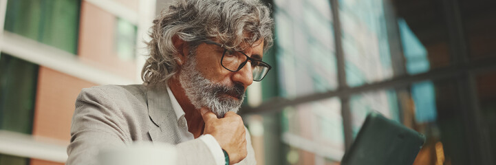 Panorama, Mature businessman with beard in eyeglasses wearing gray jacket, working on tablet, sitting in an outdoor cafe. Successful man sitting at table in cafe in nature Mobile office freelancer