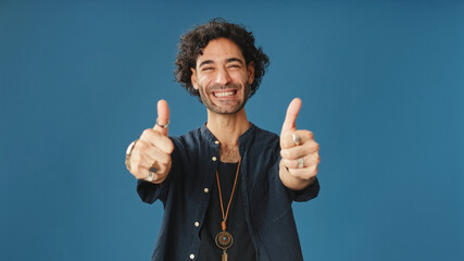 Smiling attractive man with curly hair, dressed in blue shirt, shows double thumbs up looking at...