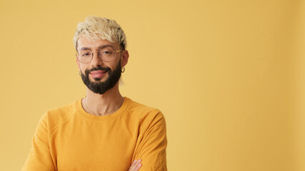 Guy with glasses, dressed in yellow T-shirt, crosses his arms and smiles, isolated on yellow...
