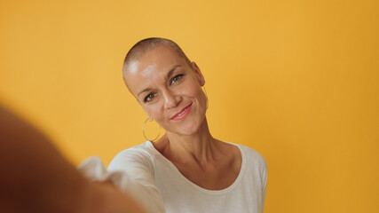Close-up, young hairless woman taking selfie looking at camera isolated on yellow background in...