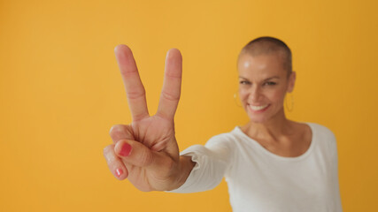 Young hairless woman, shows fingers victory gesture at camera, soft focus, isolated on yellow...