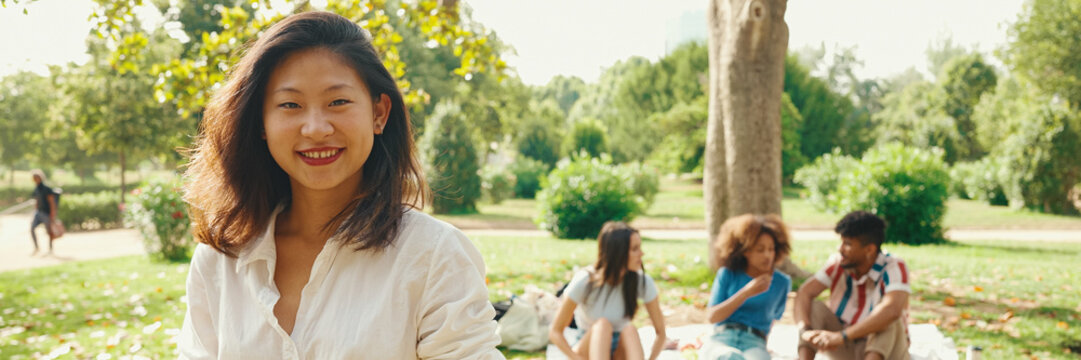 Young Asian smiling woman with long brown hair wearing white t-shirt posing for the camera in the park, Panorama. Picnic on summer day outdoors her friends sitting in distance blurred on background