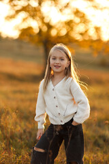 Children's day. Portrait of adorable little child caucasian girl 5-6 years smiles and looks at camera with crossed hands her hand outdoors in fall sunset lights. Leisure activity on autumn holiday.