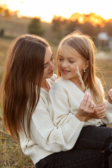 Family day, mother's day. Happy smiling young mom and adorable child daughter soft hugging, kissing and spending time together at autumn on sunset. Idyllic family having fun outdoors on fall holiday.