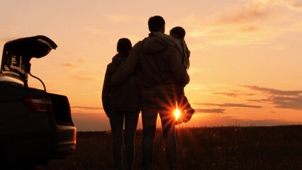 Silhouettes of happy family at sunset near car. Dad mom and girl kid watching contemplating setting sun, orange stunning summer sky. Solitude of family in nature, break from hustle and bustle of city - obrazy, fototapety, plakaty