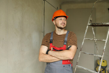 Renovation apartment. Portrait of young confident male foreman constructor repairman in safety hard hat and overalls looks at camera. Empty concrete walls, repairs house, stepladder on background.