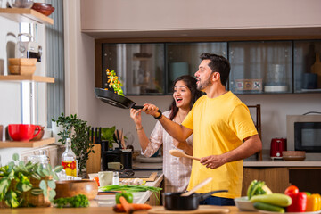 Asian Indian happy couple having fun while tossing vegetable in pan in kitchen.