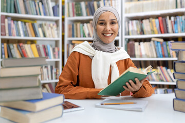 A cheerful Muslim woman in a hijab sits at a desk surrounded by piles of books in a library, smiling while holding a green book.