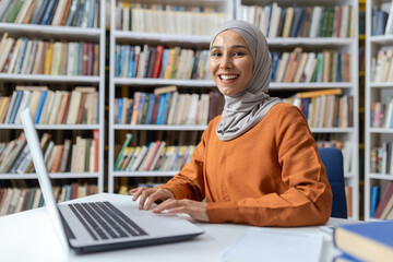 A cheerful young woman wearing a hijab sits at a library table, using her laptop among rows of...