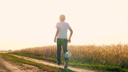 Cheerful teen boy running on dirt road at sunny sunset sunrise wheat field enjoy freedom happy...