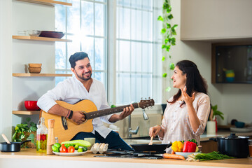 Asian Indian happy couple having fun while cooking food in kitchen, husband plays acoustic guitar.
