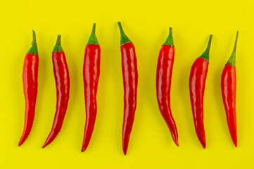 A row of red peppers are lined up on a yellow background. The peppers are all different sizes and are arranged in a straight line.