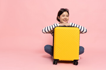 Happy Asia woman traveler sitting on floor with yellow suitcase isolated on pink background,...