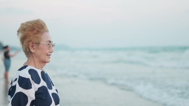 Face close up 4K with copy space of beautiful Asian elderly woman who is on the beach and looking to the sunset sky and sea with smiley face shows concept of happy, relax and enjoy retirement.