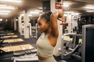 A strong black sportswoman is doing overhead triceps extension with dumbbell at gym.