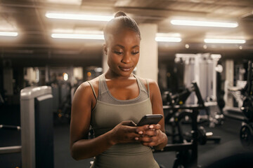 Portrait of a fit black sportswoman at gym typing messages on the phone.