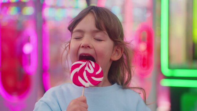 Happy smiling little girl in amusement park. Child with a large candy on a stick in her hand smiles and shows her emotions in the frame, standing against the background of burning carousels
