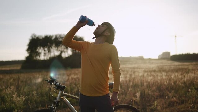 Cyclist drinks water from bottle in field during break at sunset. Relax after long bike ride in picturesque place. Caucasian man quenches thirst and replenishes level of electrolytes in hot summer.