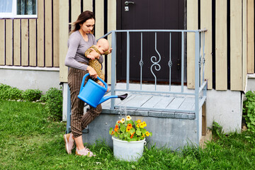 Young woman with baby in her arms waters flower bed from garden watering can.