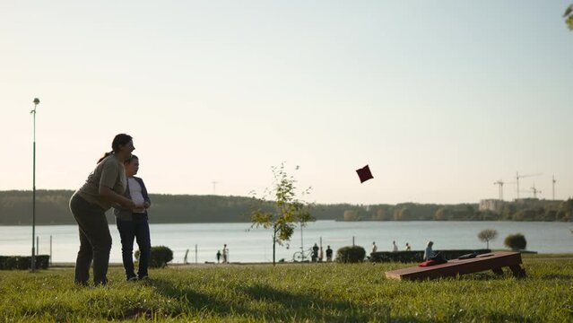 Woman and child play cornhole in park near lake. Mom and son throw bean bags into target hole while having fun outdoors in sunlight. Family picnic day with wooden game.