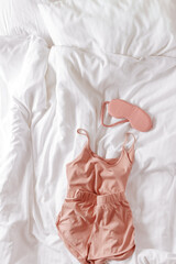 Pink female pajama and sleep eye mask on white bedclothes background. Top view comfort pyjamas for...