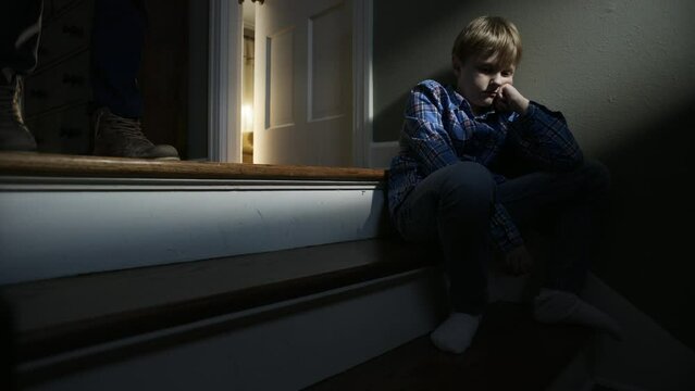 A sad boy sits on steps in the darkness while a male figure walks up behind him