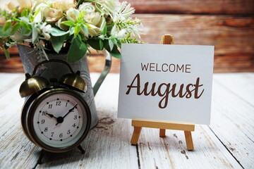 Welcome August text message written on paper card with wooden easel and alarm clock with flower in...