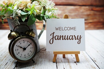 Welcome January text message written on paper card with wooden easel and alarm clock with flower in...