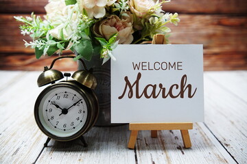 Welcome March text message written on paper card with wooden easel and alarm clock with flower in...