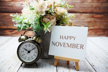 Happy November text message written on paper card with wooden easel and alarm clock with flower in...